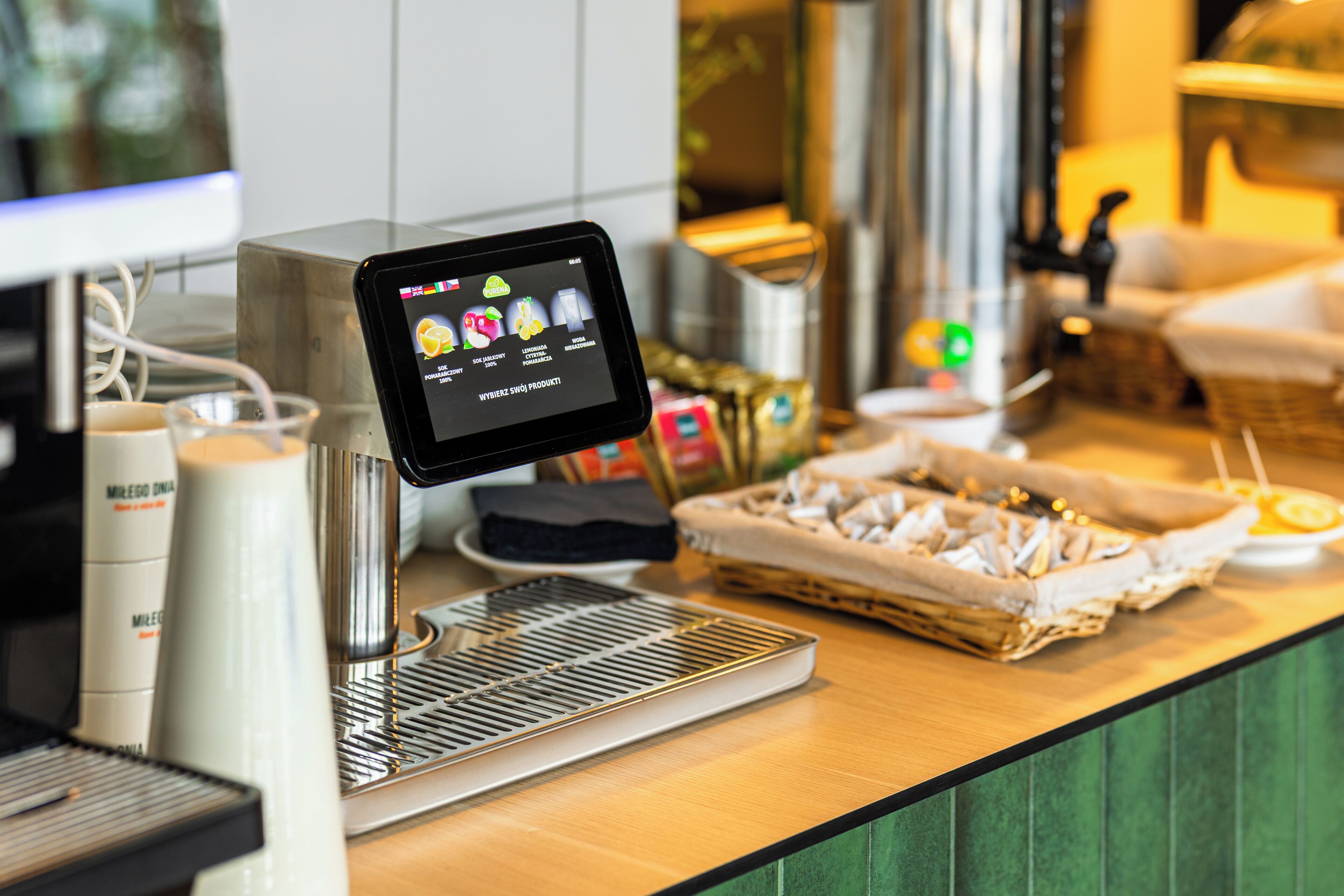 A coffee shop counter with a digital menu display, coffee machines, and a basket of pastries.