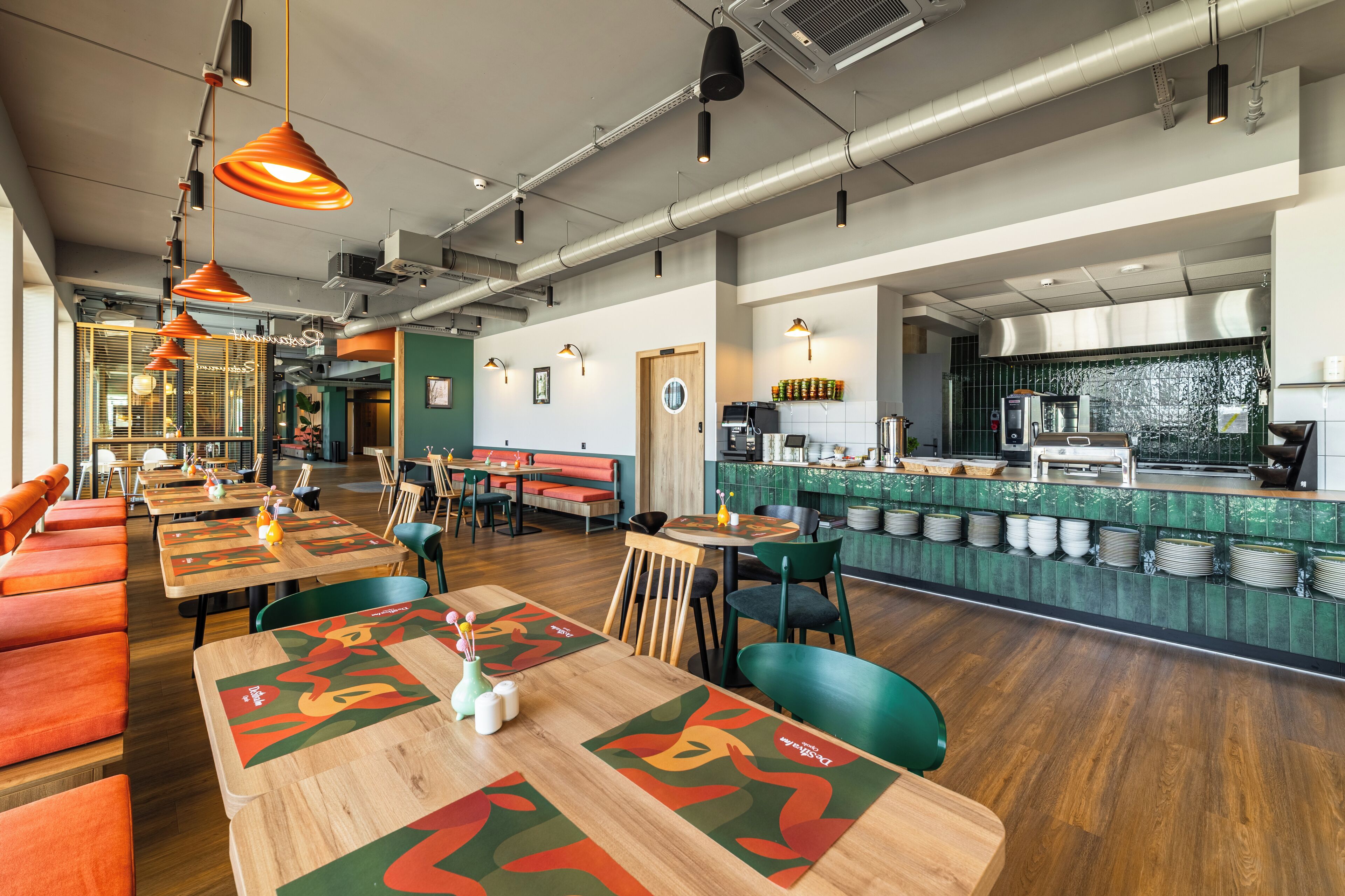 A restaurant with wooden tables, orange booths, and a kitchen area with a green backsplash.