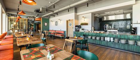 A restaurant with wooden tables, orange booths, and a kitchen area with a green backsplash.