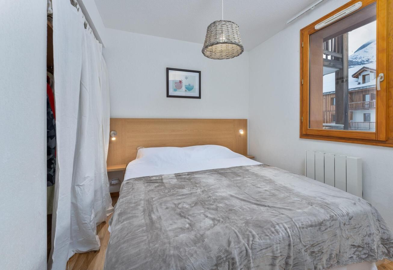 Quarto com cabeceira de madeira, colcha cinza, luminária pendente, quadro na parede e janela com vista para edifícios.