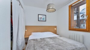 Een slaapkamer met een houten hoofdbord, een grijze spreideken, een hanglamp, een lijst met een foto aan de muur en een raam met uitzicht op gebouwen.