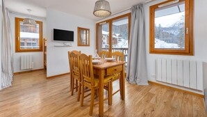 Een eethoek met een houten tafel en stoelen, een televisie aan de muur en grote ramen voorzien van gordijnen.