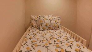 A small, single-bed room with a white bedspread featuring a lemon pattern, a white crib, and matching pillows.