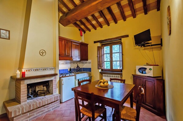 A kitchen with a fireplace, wooden cabinets, a dining table with chairs, a microwave, and a window with a view of trees.