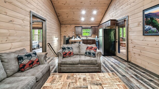 Ein gemütliches Blockhauss-Interieur mit einer Couch, einem Couchtisch und einer sichtbaren Küchenzeile durch eine geöffnete Tür.