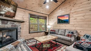 Ein gemütliches Blockhaus-Interieur mit Kamin aus Stein, Holzwänden, einem grauen Sofa mit gemusterten Kissen und einem roten und schwarzen Teppich.