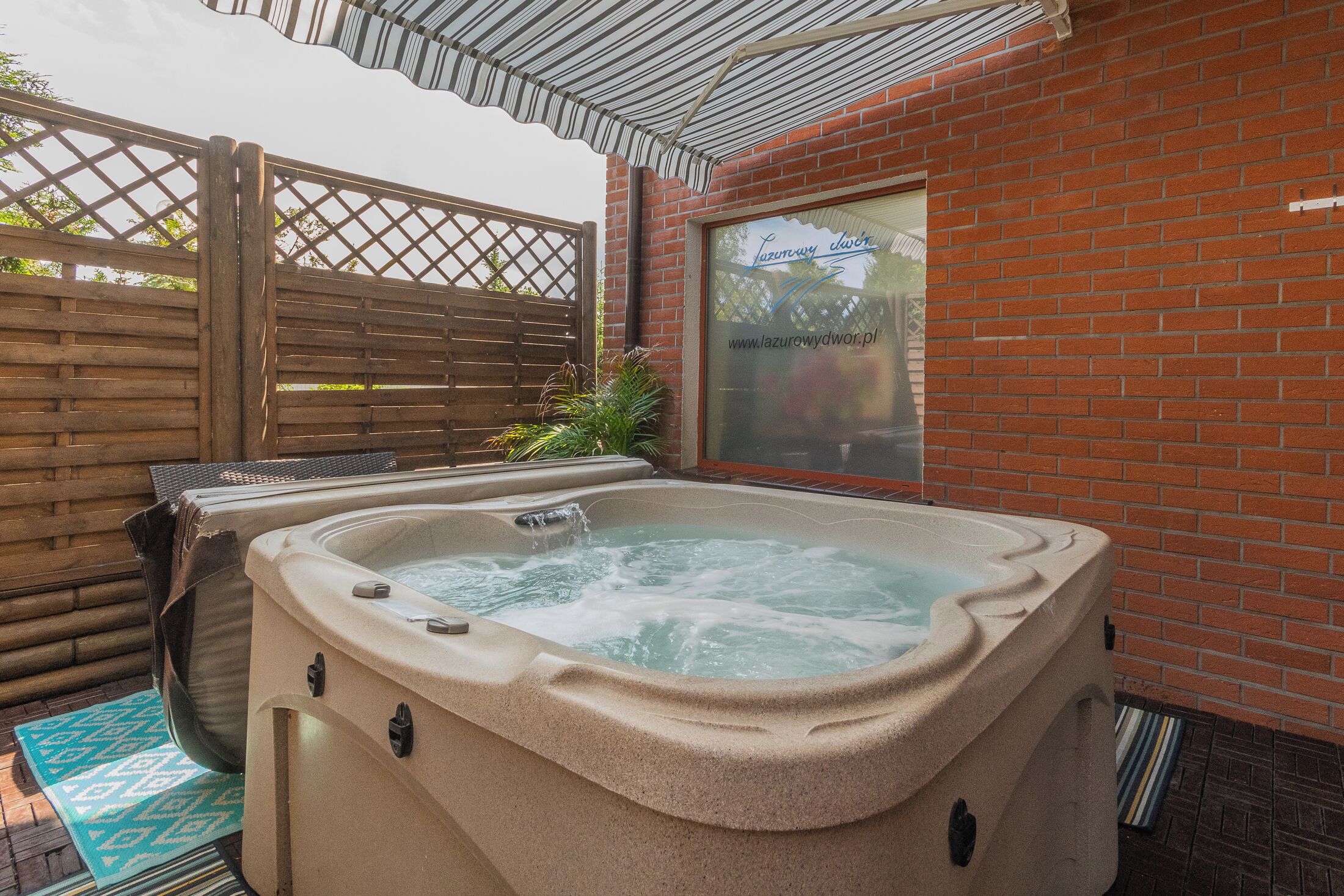 Un jacuzzi en un espacio exterior con paredes de ladrillo, cerca de una valla de madera y una puerta de cristal que muestra una dirección web.
