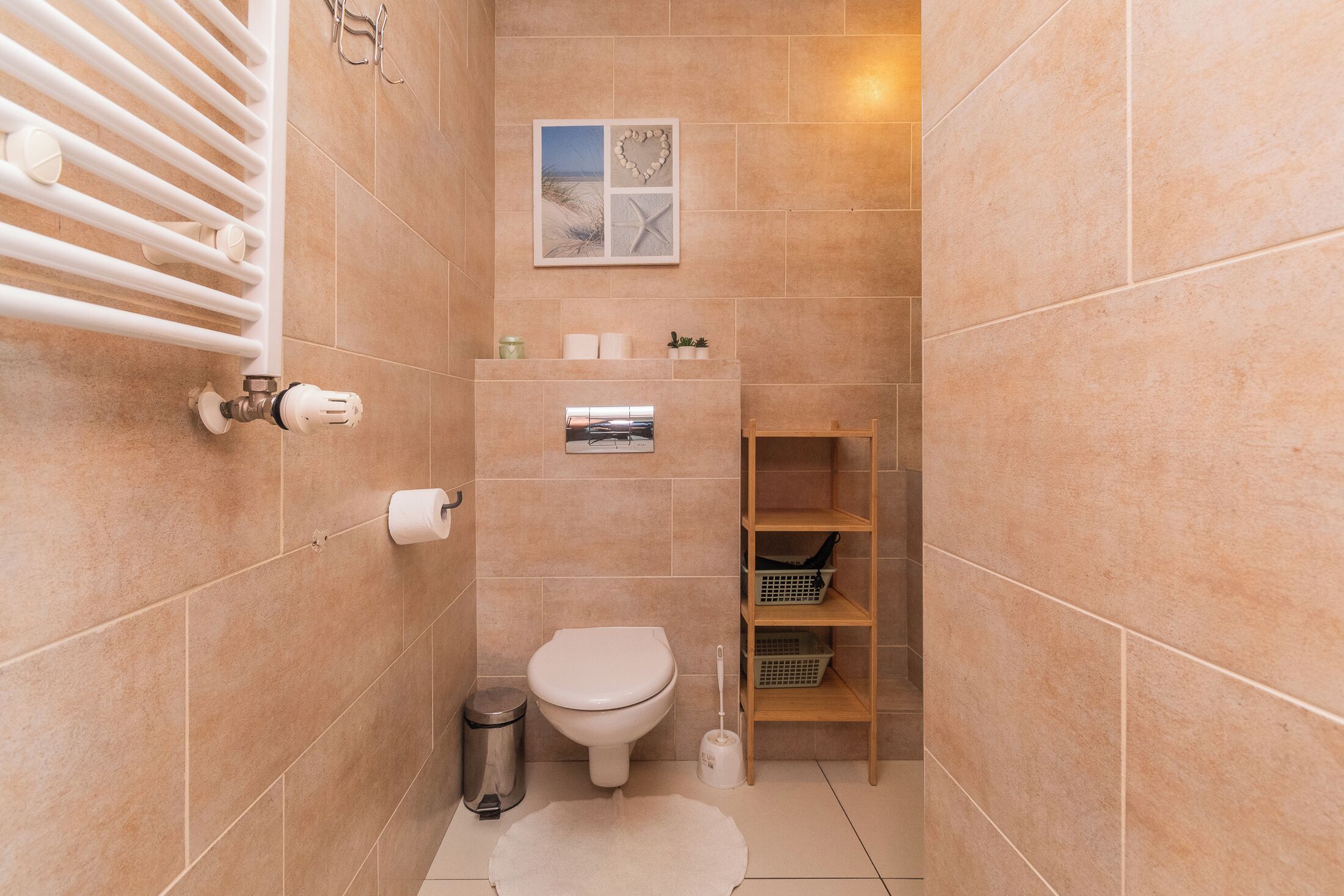 A small bathroom with beige tiles, a toilet, a radiator, a shelf with baskets, and a framed picture on the wall.