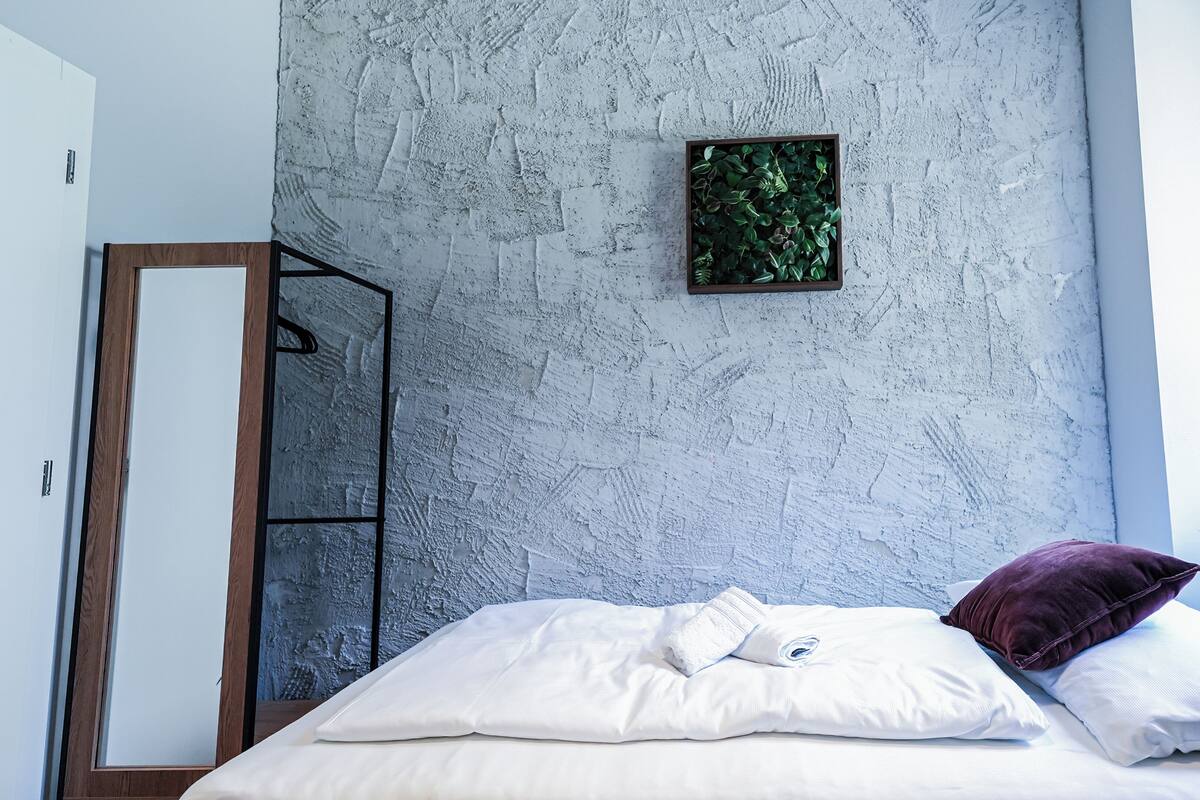 A bedroom with a bed, white pillows, a purple pillow, a framed plant, and a wardrobe.