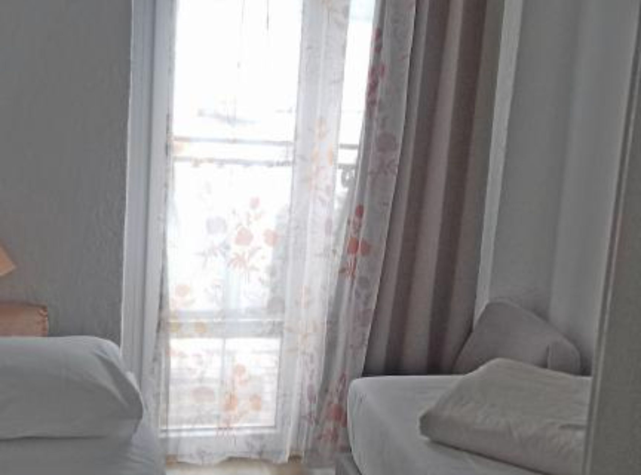 A room with two beds, a window with floral curtains, and a lamp on a bedside table.