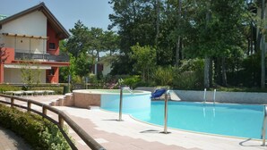 A swimming pool area with a wooden deck, lounge chairs, and a building with a balcony.