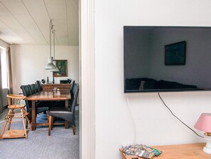 A room with a dining table, chairs, a television mounted on the wall, and a lamp on a side table.