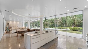 A modern kitchen with a large island, a dining area with a table and chairs, and large windows offering a view of a garden and pool. - Floral City Oasis / Pool, BBQ Area & Forest View (Floral City)