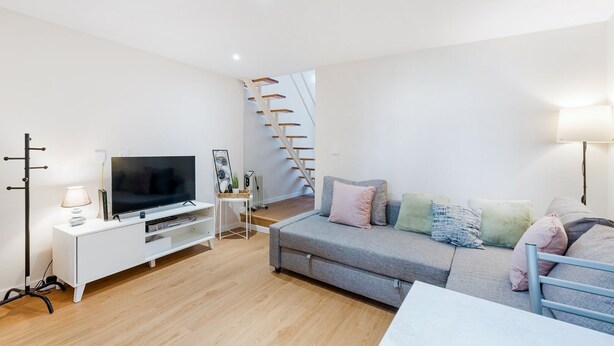 Un salon moderne doté d’un téléviseur à écran plat, d’un meuble de rangement blanc, d’un canapé gris agrémenté de coussins colorés, d’un plancher en bois et d’un escalier.