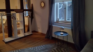 A room with a wooden floor, a radiator, a window with curtains, a round mirror, and a small table with a lamp.