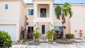 A two-story house with a front porch, a garage, and a well-maintained garden with palm trees and various plants. - Tangerine Sunsets Pomelo East (Nassau Bahamas)