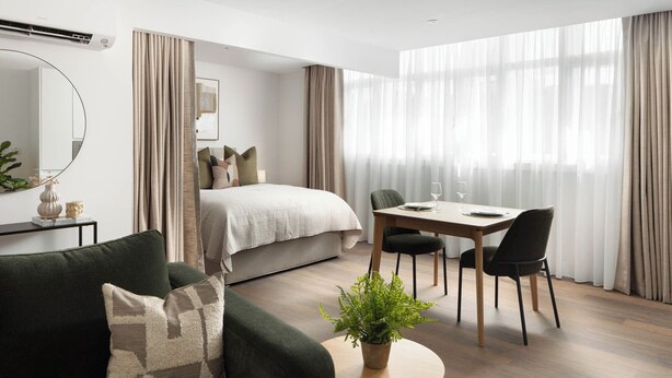 A modern hotel room with a bed, a dining table with chairs, a green sofa, a round wooden table with a plant, and a large window with sheer curtains. - Urban Rest Dublin Central (Dublin)