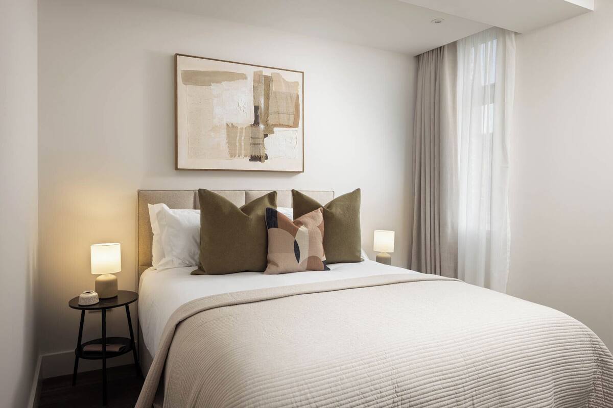 Une chambre d’hôtel avec un grand lit, deux tables de chevet avec des lampes, un tableau abstrait encadré au mur et une fenêtre avec des rideaux.