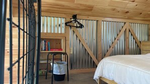 A bedroom with a bed, a small stool, a shelf with books, and a hanging light fixture. - Quiet 2-bedroom country cabin with scenic views 3 miles from Norwich NY. (Norwich)