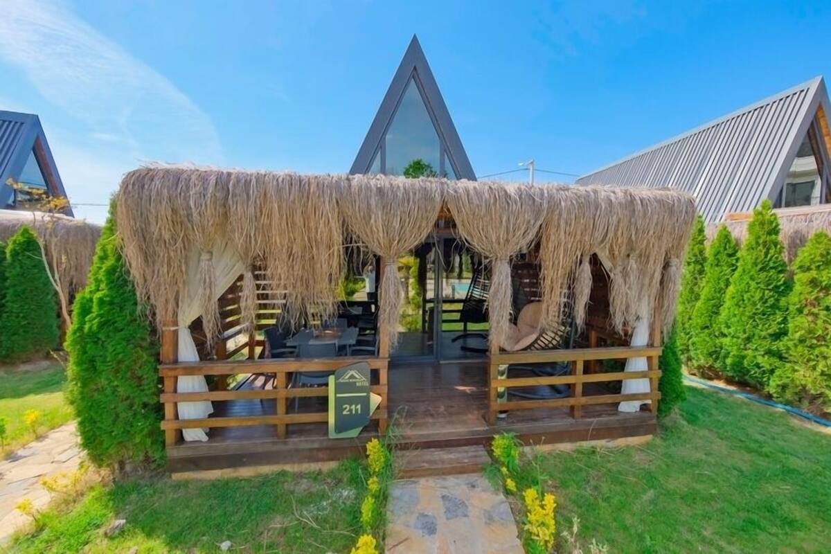 A thatched-roof outdoor seating area with a wooden deck, a sign indicating room number 211, and a modern building in the background.