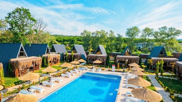 Ein Swimmingpool mit Liegestühlen und Sonnenschirmen, umgeben von Hütten mit Reetdächern.