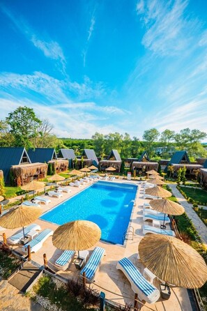 Ein Swimmingpool mit Liegestühlen und Sonnenschirmen, umgeben von Hütten mit Reetdächern.