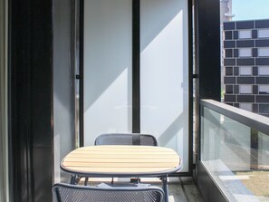 Un balcón con mesa y sillas, un ventanal de vidrio y vistas a un edificio con fachada enrejada.