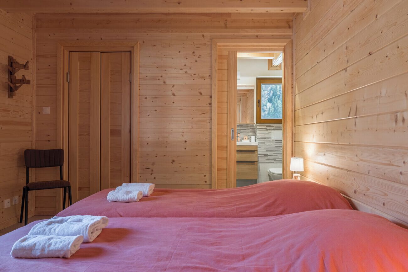 A wooden cabin room with two beds, a chair, and a bathroom visible through an open door.