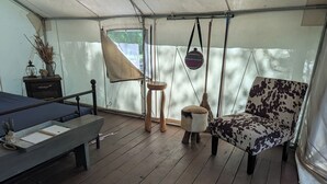 Ein Zeltraum mit einem Bett, einem kleinen Tisch mit einem Notizbuch, einem Stuhl, einer aufgehängten Tasche und einem Besen.