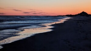 Ein Strand bei Sonnenuntergang mit Wellen, einer entfernten Hügelkette und Fußabdrücken im Sand.