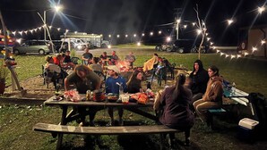 Eine Gruppe von Menschen versammelt sich abends draußen an einem Tisch mit Essen und Getränken, darüber hängen Lichterketten.