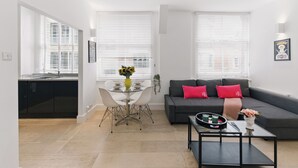 Una sala de estar moderna con un sofá gris, una mesa de centro y una mesa de comedor con sillas. Hay dos ventanas con persianas, un área de cocina y una planta pequeña en el alféizar.