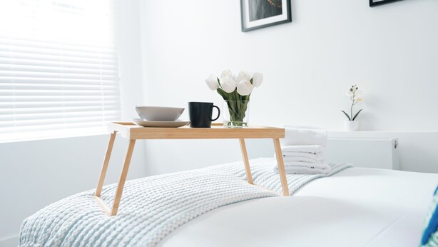 Una cama bien hecha con una manta blanca y azul, una pequeña mesa de madera con un cuenco, una taza negra y un jarrón con flores blancas.
