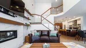 A modern living room with a fireplace, a brown leather sofa, a wooden coffee table, and a kitchen area with wooden cabinets and bar stools.