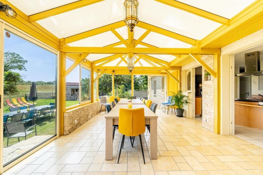 Un espacio comedor al aire libre con techo, una mesa larga y sillas amarillas, ventanas grandes y una pared de piedra.