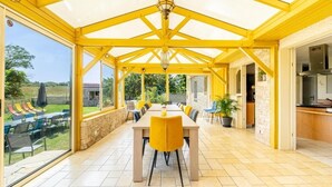 Un espacio comedor al aire libre con techo, una mesa larga y sillas amarillas, ventanas grandes y una pared de piedra.
