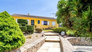 Una casa amarilla con techo de tejas, rodeada de setos cuidados y un camino de piedra que conduce a la entrada.