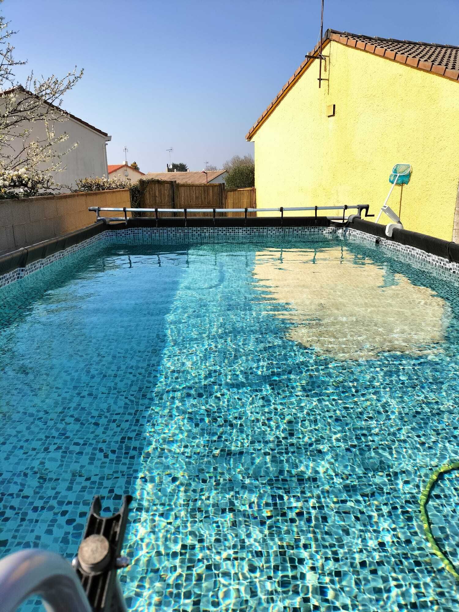 A clear swimming pool with a tiled bottom, surrounded by a wooden fence and a yellow building.