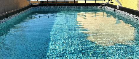 A clear swimming pool with a tiled bottom, surrounded by a wooden fence and a yellow building.