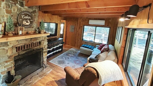 Ein gemütliches Blockhaus-Interieur mit Kamin aus Stein, Holzwänden, einer Couch und einem großen Fenster mit Aussicht.