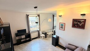 A living room with a sofa, a TV, a dining area with chairs, a table, and a window with curtains. - Vista Mare Antico [Mura delle Grazie] (Genoa)