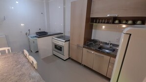 A compact kitchen with a stove, sink, and a small refrigerator. There is a dining table with chairs and a white cabinet.