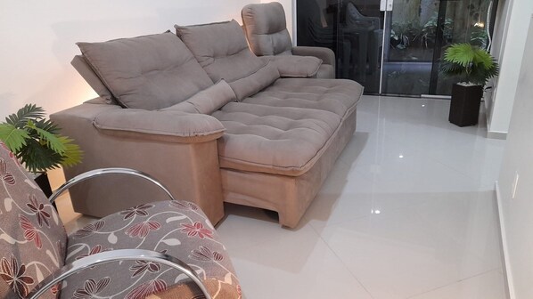 A modern living room with a sectional sofa, a patterned armchair, and a potted plant. - Apto 103 Ed Dambros bem localizado (Piratuba)