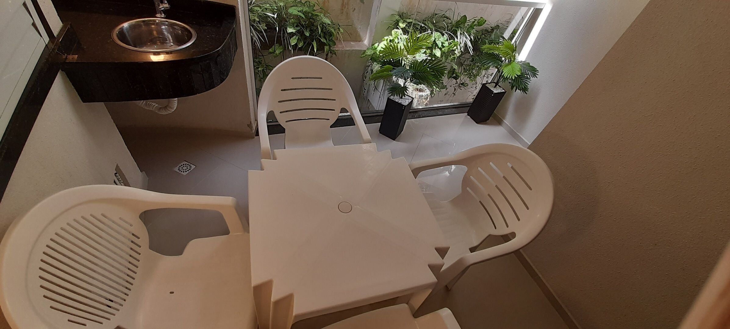 Un petit coin repas avec une table blanche et des chaises en plastique, un plan de travail noir avec un évier, et des plantes en pot en arrière-plan.