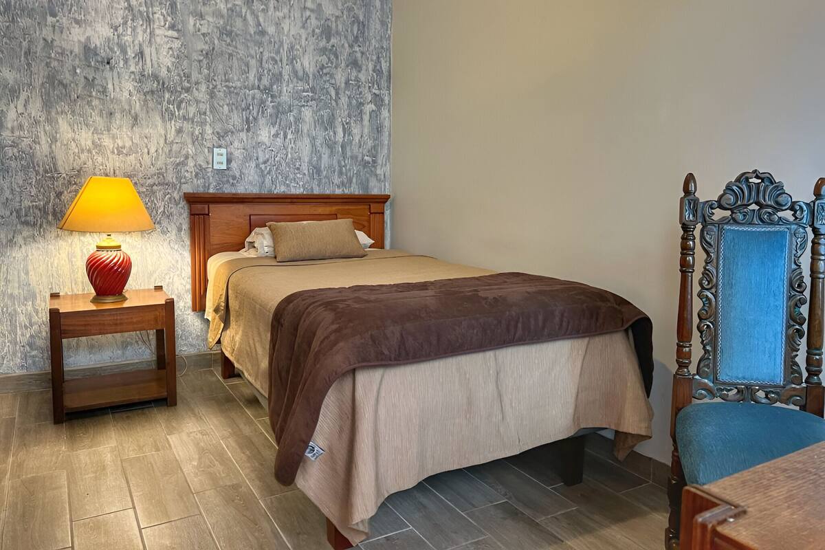 A neatly made bed with a wooden headboard, a bedside table with a lamp, and a chair with a blue cushion.