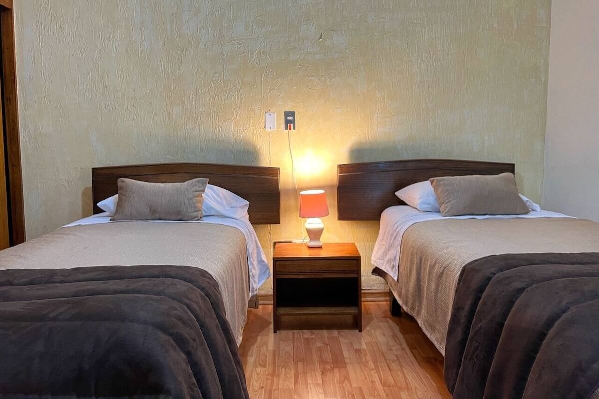 A room with two beds, each with a wooden headboard, a nightstand with a lamp, and a wall socket.