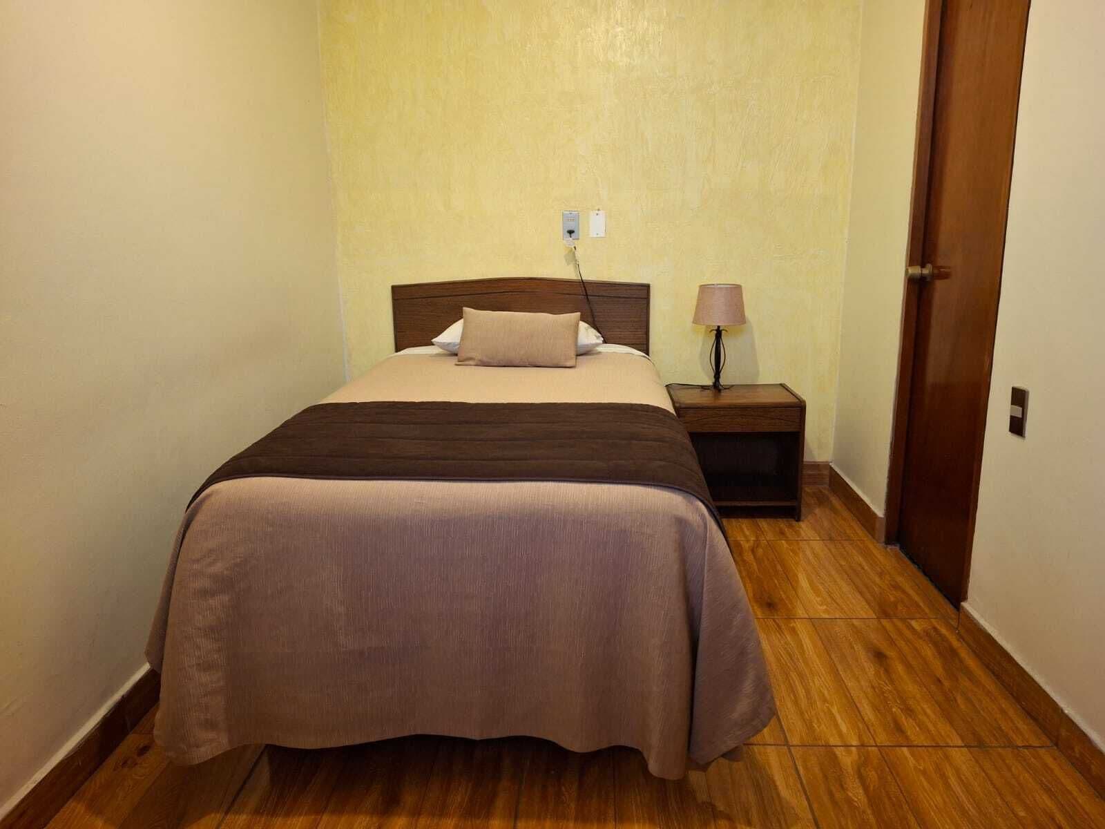 Quarto de hotel com uma cama, mesa de cabeceira, abajur e porta.
