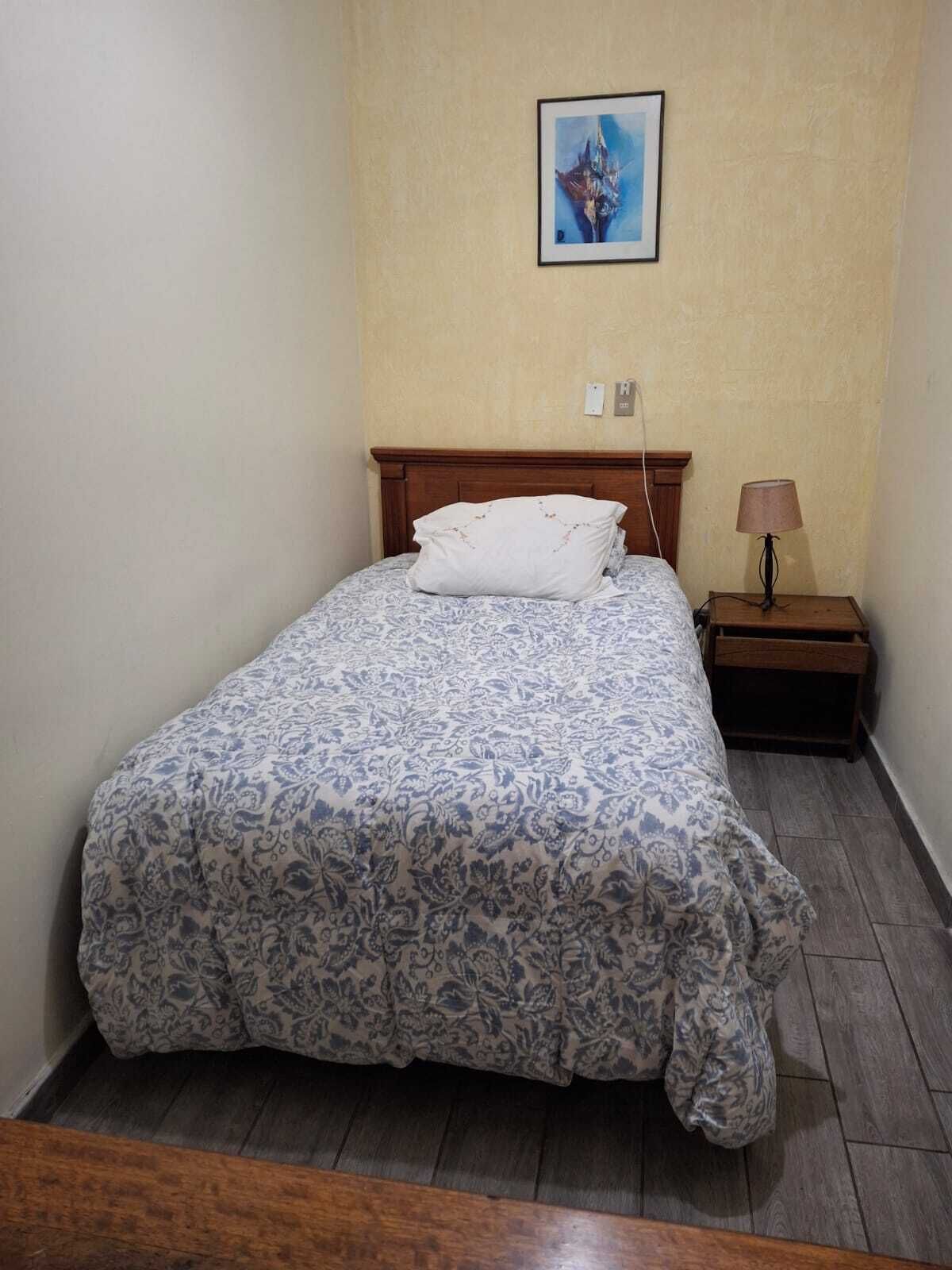 A single bed with a floral-patterned bedspread, a wooden headboard, a bedside table with a lamp, and a framed picture on the wall.