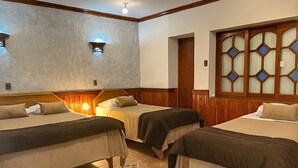 Une chambre d’hôtel avec trois lits simples, des têtes de lit en bois, un sol carrelé et une fenêtre décorative avec des vitres en verre bleu.
