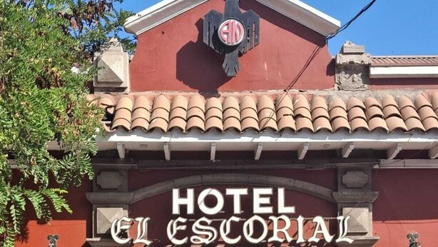 L’entrée d’un hôtel, avec une façade rouge, un toit recouvert de tuiles et une enseigne indiquant « HÔTEL ESCORIAL ».